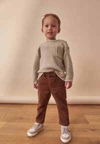 Valkoinen harmaa neulepusero, jossa on tasku, ruskeat samettilautaset ja beige tennarit. Asusteessa on kontrastisia tekstuureja ja yksinkertainen muotoilu.