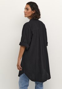 Chemise noire à manches roulées, en tissu texturé. Dotée d'un pli au dos et d'un ourlet arrondi, associée à un jean bleu.
