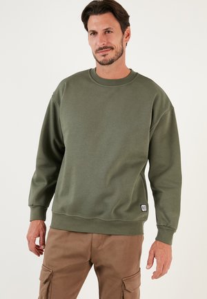Sudadera de algodón verde oliva con escote redondo, puños acanalados y un pequeño parche de logo en el dobladillo. Tejido texturizado y corte relajado.