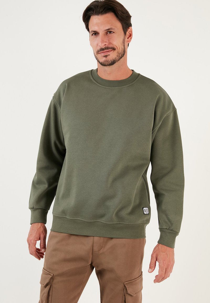 Sudadera de algodón verde oliva con escote redondo, puños acanalados y un pequeño parche de logo en el dobladillo. Tejido texturizado y corte relajado.