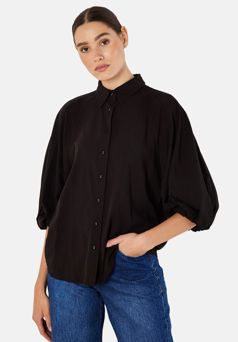 Schwarzes Hemd mit strukturiertem Kragen, gepufften Ärmeln und glatter Textur. Kombiniert mit blauen Jeans für einen kontrastierenden Look.