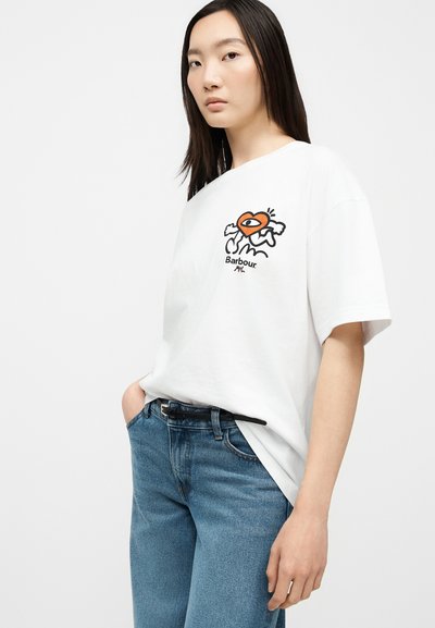 Femme portant un T-shirt blanc oversize avec un cœur orange graphique et un texte noir, associé à un jean bleu taille haute et une ceinture noire.