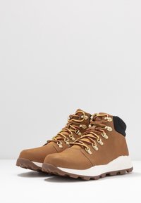 Bruna vandringskängor med hög ankel, med guldfärgade ögla, röda och gula snören samt en vit strukturerad sula. Slät ovandel.