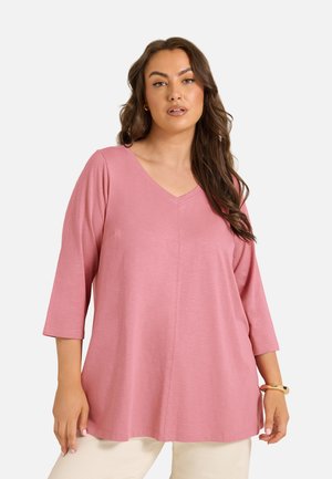 Femme aux longs cheveux bruns porte un haut rose ample à col en V avec des manches trois-quarts et un pantalon beige sur fond uni.