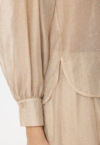 Beige lightweight blouse with sheer fabric, featuring silver sequins and a rounded hem. Long sleeves gathered at the cuffs with button closure.