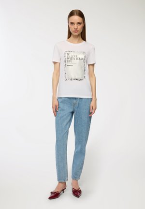 T-shirt bianca a maniche corte con stampa grafica che recita "AMATI LA VITA CHE HAI", abbinata a jeans azzurri a gamba dritta e scarpe con tacco rosse.