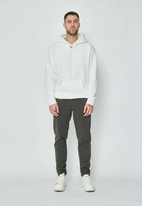 Jeune homme debout, faisant face à l'avant, portant un sweat à capuche blanc, un pantalon sombre, des baskets blanches et des bagues aux deux mains.