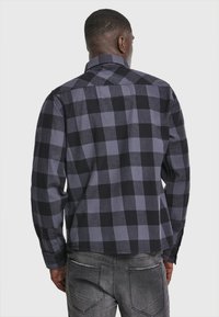 Camisa de franela a cuadros en negro y gris, de manga larga, con cuello y dobladillo recto, que presenta una tela texturizada y puños abotonados.