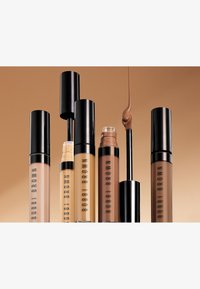 Fem bobbi brown concealer rør med svarte lokk, i varierende nyanser fra lys beige til mørk brun, en applikator stikker ut fra ett av rørene.
