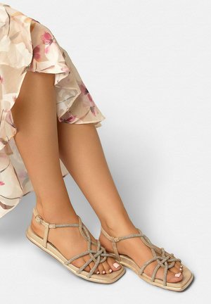 Vrouwelijke benen met beige platte sandalen met bandjes en rhinestones, gecombineerd met een jurk met bloemenprint en gerimpelde zoom tegen een effen achtergrond.