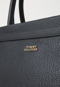 Sort tekstureret læder tote taske med guld "TOMMY HILFIGER" logo, som har glatte håndtag og et elegant, minimalistisk design.