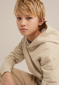 Beige Kapuzenpullover mit einer Fronttasche, gerippten Bündchen und einem Kordelzug. Der Stoff wirkt weich mit einer glatten Textur und wird lässig mit passenden Hosen getragen.