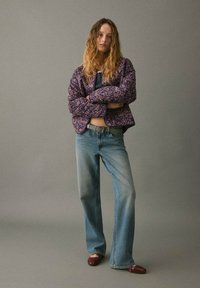 Giacca trapuntata con motivo floreale viola, top blu navy, jeans svasati azzurri chiari e scarpe bordeaux. Tessuto textured con contrasti accentuati.