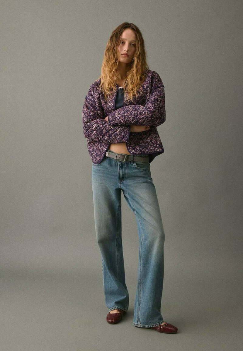 Giacca trapuntata con motivo floreale viola, top blu navy, jeans svasati azzurri chiari e scarpe bordeaux. Tessuto textured con contrasti accentuati.