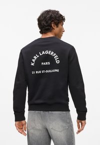 Sudadera negra con cuello redondo, puños y dobladillo de canalé. Presenta texto blanco en la espalda: "KARL LAGERFELD PARIS 21 RUE ST-GUILLAUME."
