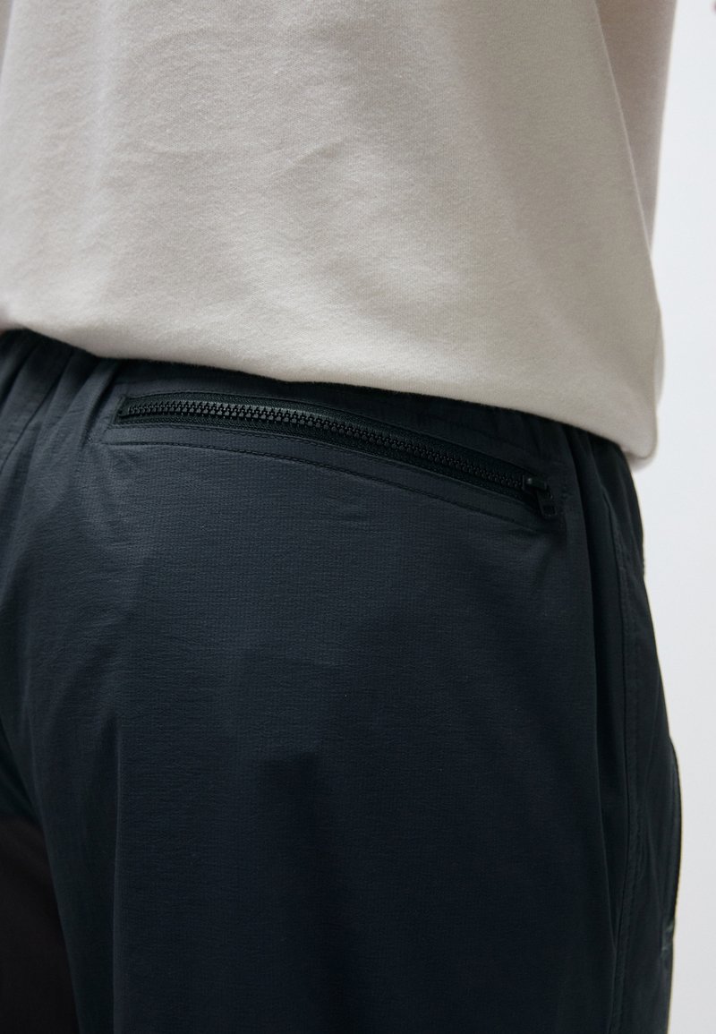 Svarta lätta byxor med en texturerad yta, försedd med en ficka med dragkedja på baksidan, i kombination med en ljusgrå topp.