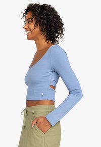 Lys blå langærmet crop top i ribbet stof med en rund halsudskæring og sideudskæringer, parret med olivengrønne bukser med snoretræk.