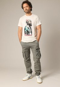 T-shirt en coton blanc avec un imprimé graphique d'un homme barbu portant des lunettes, associé à un pantalon cargo gris clair et des baskets blanches avec des accents bleus.