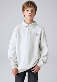 Chlapec se světlými hnědými vlasy, oblečený v bílém polokošili s dlouhým rukávem a nápisem „ALLSAINTS“, stojící před jednolitým šedým pozadím.