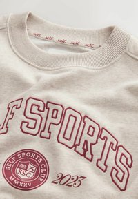 Lysebeige sweatshirt med ribbet krave, med vinrød broderet tekst "SELF SPORTS" og et cirkulært logo med "2023" og "SSC."