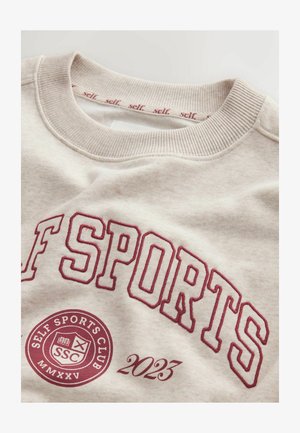 Helebeež vest, millel on soonikkrae, kaunistatud tumepunase tikitud tekstiga "SELF SPORTS" ja ringikujulise logoga, mis sisaldab numbrit "2023" ja "SSC".