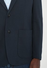 Blazer azul marino hecho de tejido texturizado, con solapa con muesca, tres botones negros y un solo bolsillo en el pecho. Estilizado con vaqueros.