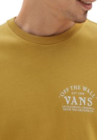 Camiseta de algodón amarilla mostaza con un cuello redondo acanalado, que presenta un logotipo gráfico en blanco que dice "OFF THE WALL" y "VANS."