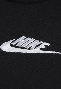 Il tessuto nero presenta un logo Nike bianco ricamato, che mostra un design curvo con un aspetto aerodinamico e una scritta distintiva.