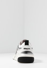 Zapatilla blanca y plateada con detalle de malla, forma redondeada, suela de goma negra y pestaña de talón con marca. Texturas y acentos en contraste notables.