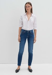 Witte button-up shirt met opgerolde mouwen, gecombineerd met blauwe skinny jeans en zwarte ballerina's, tegen een simpele, lichte achtergrond.