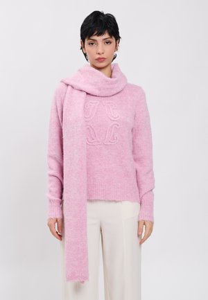 Frau trägt einen hellrosa gestrickten Pullover mit einem passenden Schal, der über eine Schulter gelegt ist, kombiniert mit weißen Hosen, stehend vor einem einfarbigen Hintergrund.