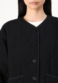Black quilted jacket featuring a rounded collar, five black buttons, and side pockets with a textured wave pattern.