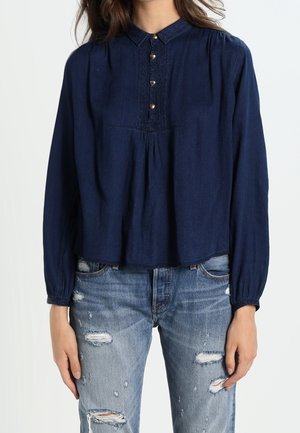 Vrouw draagt een navy blauwe blouse met lange mouwen en knopen aan de voorkant en lichtblauwe gescheurde jeans, staat met ontspannen armen langs haar zij.