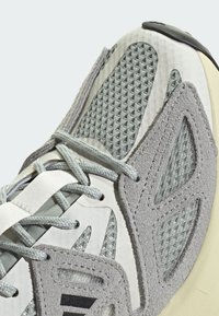 Gray athletic shoe featuring mesh and suede materials, textured accents, flat laces, and a smooth rubber sole. Distinctive side detailing.
