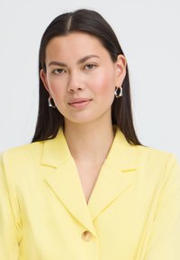 Jeune femme aux longs cheveux bruns portant des boucles d'oreilles argentées en forme de créoles et un blazer jaune, regardant droit devant avec une expression neutre.