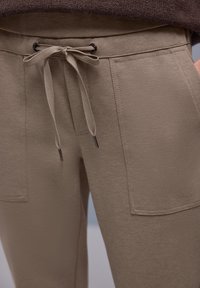 Beige broekstof met een textuurafwerking, voorzien van een verstelbare taille met trekkoord, twee voorzakken en donkere metalen accenten.