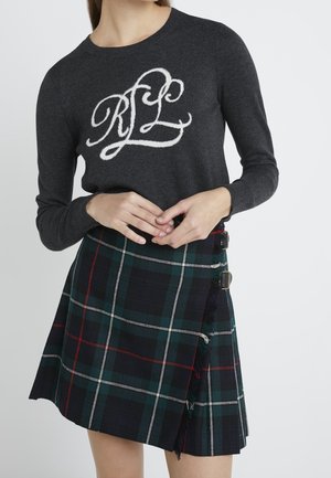 Kvinde iført mørkegrå sweater med hvide "RL" bogstaver og grøn, blå og rød ternet omslagsnederdel med spænde detaljer.