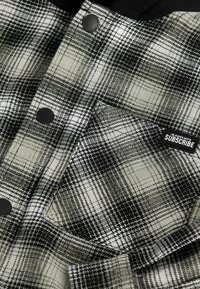 Chemise à carreaux noir et blanc avec boutons-pression noirs, poche poitrine et étiquette rectangulaire "SUB5CRIBE" sur la poche.