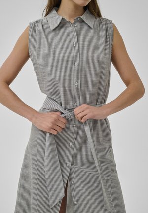 Mouwloze grijze jurk met witte verticale strepen, knoopsluiting aan de voorkant, kraag en een getailleerde taille, gedragen door een persoon die de riem verstelt.