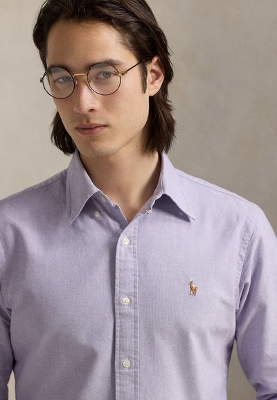 Ανοιχτό μωβ πουκάμισο από βαμβάκι, με κλασικό γιακά και κεντημένο λογότυπο στο στήθος. Λείο ύφασμα και εφαρμοστή γραμμή.