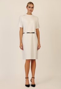 Cremefarbenes Kurzarmkleid mit hochgeschlossenem Ausschnitt, tailliert mit einem schwarzen Gürtel mit goldenen Akzenten, kombiniert mit schwarzen High Heels.