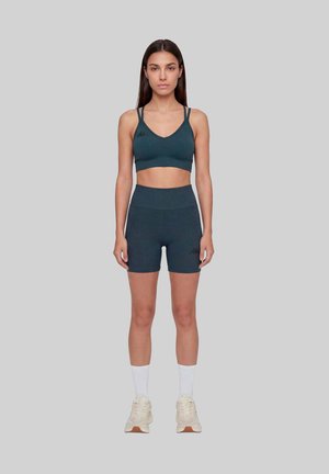 Femme debout face devant portant un soutien-gorge de sport bleu sarcelle foncé et un short de cycliste taille haute assorti, chaussettes blanches et baskets crème sur fond gris.