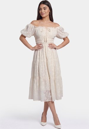 Vrouw poseert in een licht beige off-shoulder midi-jurk met pofmouwen en witte hoge hakken tegen een effen achtergrond.