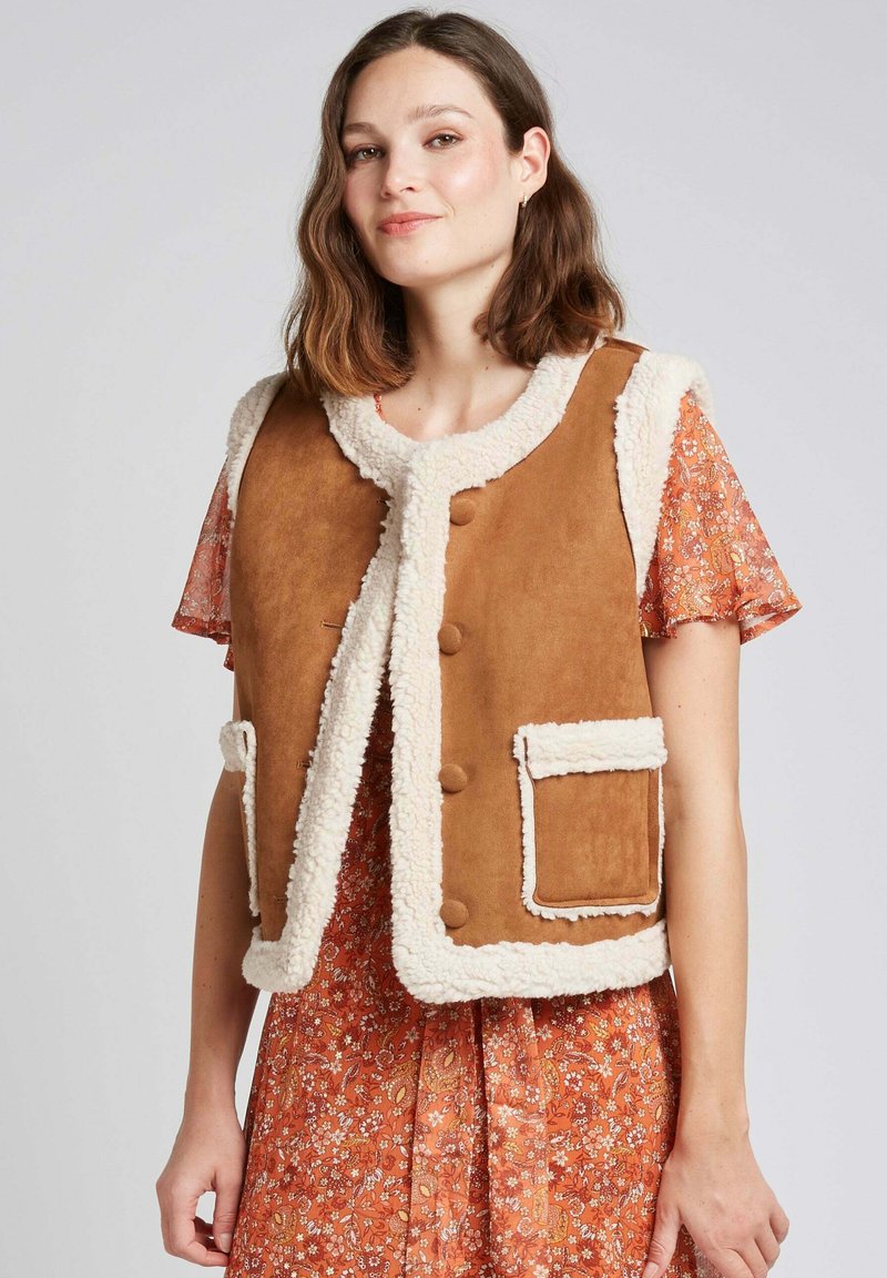 Gilet en suède marron clair avec des bordures en fausse fourrure crème ; présente un col rond, des boutons sur le devant et deux poches. Porté par-dessus une robe à motif floral.