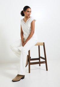 Haut blanc à col en V avec bord en dentelle, associé à un pantalon blanc évasé et des bottines beige à la cheville. Le décor comprend un tabouret en bois.