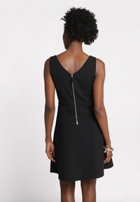 Femme avec des cheveux bouclés portant une robe noire sans manches avec une fermeture éclair au dos et des bracelets au poignet droit, se tenant devant un fond uni.