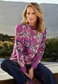 Eine Frau mit blonden Haaren sitzt im Schneidersitz auf einem Holzdeck. Sie trägt ein lila Blusenoberteil mit Blumenmuster und dunkelblaue Jeans und ist im Freien.