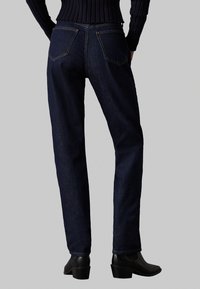 Jeans de mezclilla azul oscuro con corte recto, que cuentan con bolsillos traseros y una textura suave. Se llevan con botas negras de tacón.