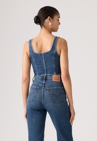 Γυναίκα με μαλλιά κομμένα κότσο που φοράει αμάνικο τζιν τοπ και ψηλόμεσα μπλε τζιν, στέκεται με την πλάτη γυρισμένη σε απλό φόντο.