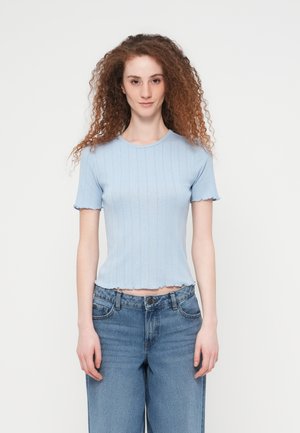 Kvinne med krøllete rødt hår iført en lyseblå kortermet ribbestrikket topp og høytlivs blå jeans, stående foran en hvit bakgrunn.